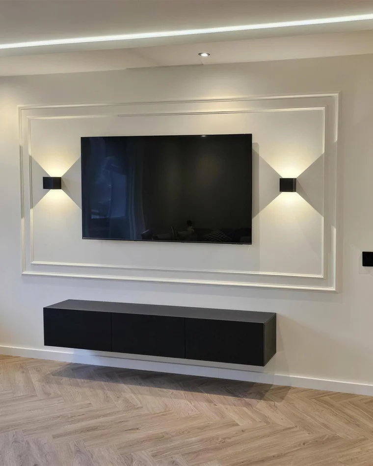 Tv-wand met witte sierlijsten, wandlampen en een zwevend tv-meubel boven een visgraatvloer